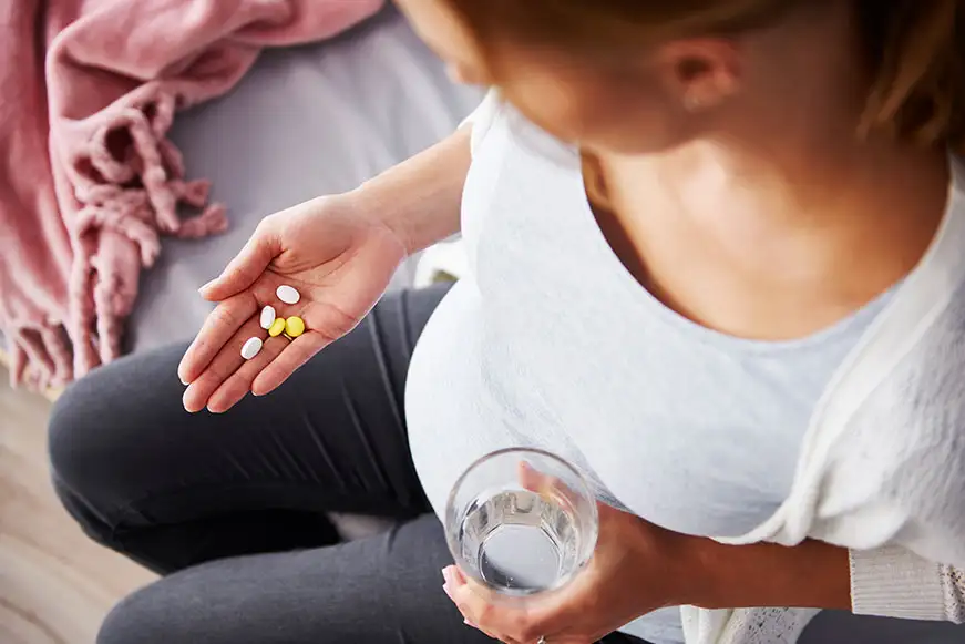 Top down view of a pregnant woman sitting on a bed, with a handful of pills in one hand and a glass of a water in the other