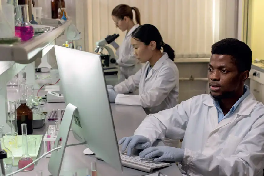 Three scientists are working in a lab, one on a microscope, one with a pipette and the final one works on a laptop 